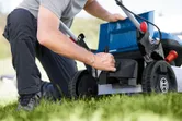 A person adjusts the compartment of a cordless lawn mower on a grassy lawn.