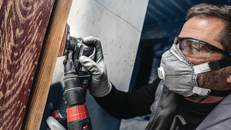 Person wearing safety equipment sands a wooden surface with a power tool.