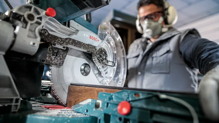 Person wearing safety equipment uses a circular saw to cut a wooden plank.