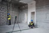 Two people wearing safety equipment align walls using a laser leveling tool in a building under construction.