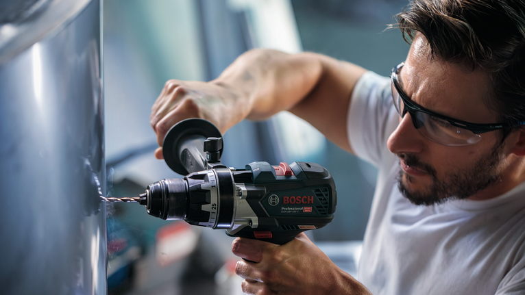 A person wearing safety equipment drills into metal with a power tool.