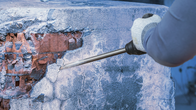 Worker wearing safety equipment uses a demolition tool to break concrete from a wall.