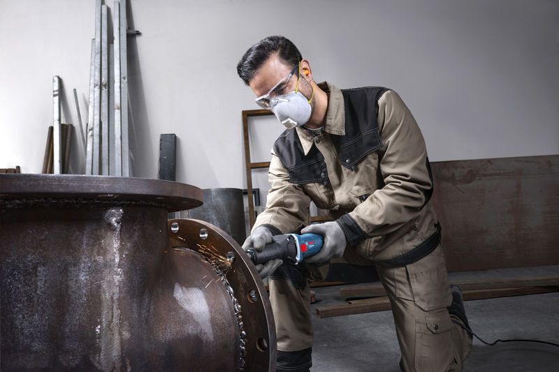 A person wearing safety equipment grinds the edge of a large metal pipe.