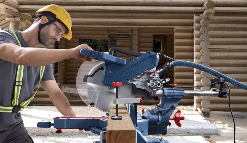 A person wearing safety equipment operates a mitre saw to cut wood at a construction site.