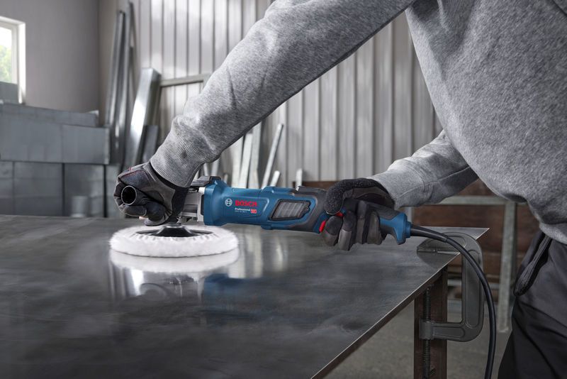 A person wearing safety equipment polishes a metal sheet with a blue power tool.