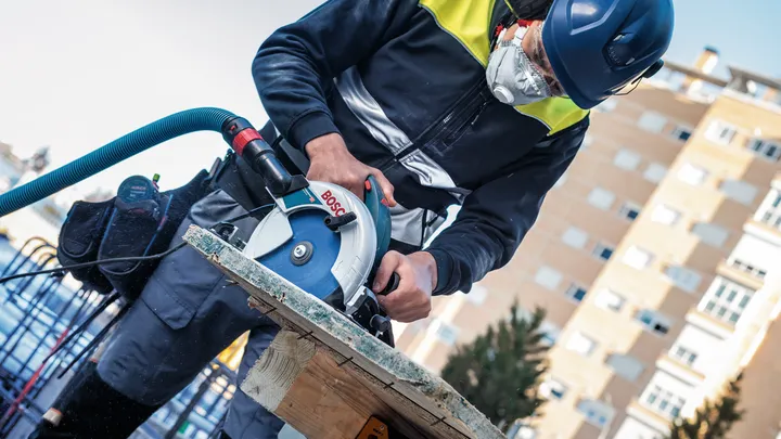 Person wearing safety equipment cuts wood with a circular saw at a construction site.