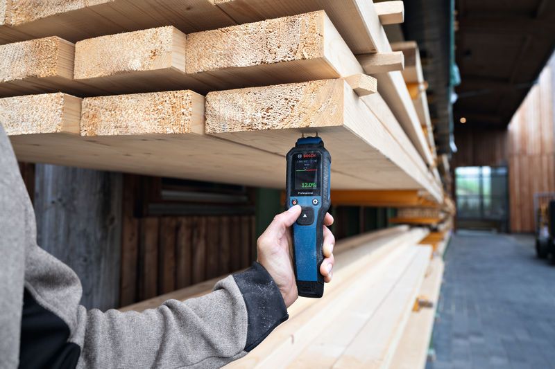 A person checks stacked timber moisture using a material moisture meter outdoors.