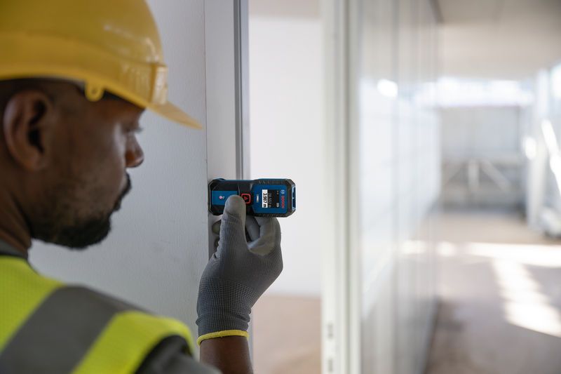 A person wearing safety equipment uses a laser measure against a door frame.