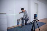 A person kneels marking a wall guided by a laser leveling tool on a tripod.