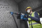 A person wearing safety equipment uses a demolition hammer on a concrete wall.