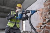 A person wearing safety equipment uses a demolition hammer with SDS max to break a brick wall.