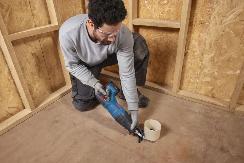 A person wearing safety equipment cuts a pipe on the floor using a cordless reciprocating saw.