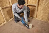 A person wearing safety equipment cuts a pipe on the floor using a cordless reciprocating saw.