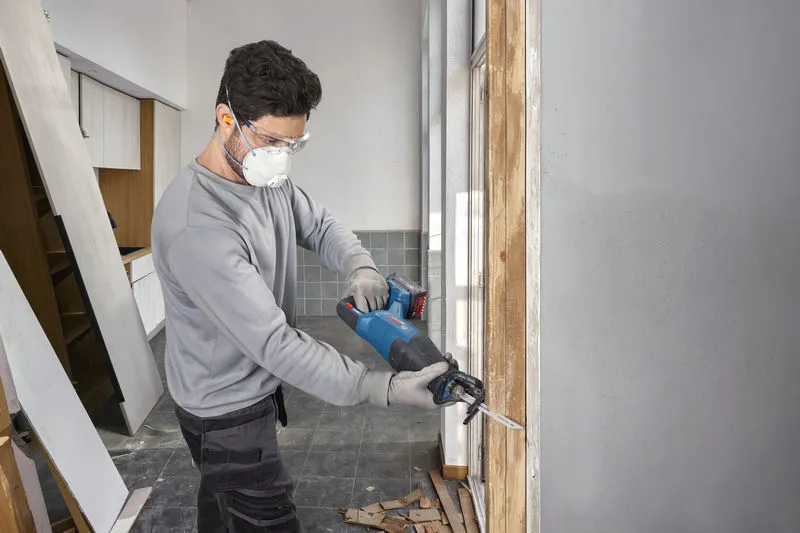 A person wearing safety equipment uses a cordless reciprocating saw to cut a wooden frame.