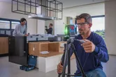 Worker wearing safety equipment adjusts laser leveling tool on a tripod in a modern kitchen.