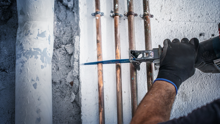 Person wearing safety equipment cuts wall near pipes with a reciprocating saw.