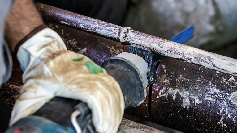 Person wearing safety equipment cuts a rusted metal pipe with a reciprocating saw.