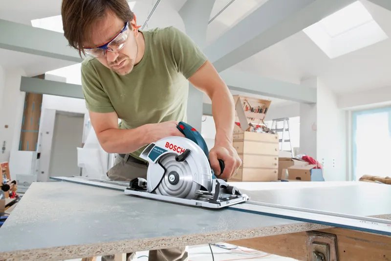 A person wearing safety equipment cuts a wood board with a circular saw.