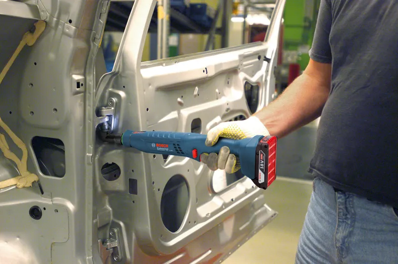 Uma pessoa que usa equipamento de segurança utiliza uma ferramenta sem fios para prender uma dobradiça dentro da porta de um carro.