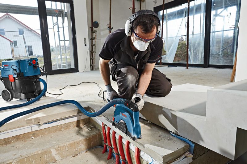 Uma pessoa que utiliza equipamento de segurança corta betão junto a tubagens expostas com uma fresadora de parede.