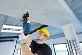 Uma pessoa que utilize equipamento de segurança perfura um teto de betão com um martelo rotativo.