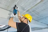Uma pessoa que utiliza equipamento de segurança utiliza um martelo rotativo para perfurar um teto de betão.