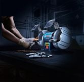 Uma pessoa que utilize equipamento de segurança afia uma broca com uma retificadora de bancada de roda dupla.