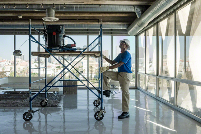 Pessoa que usa equipamento de segurança está ao lado de um andaime móvel no interior de um edifício moderno.