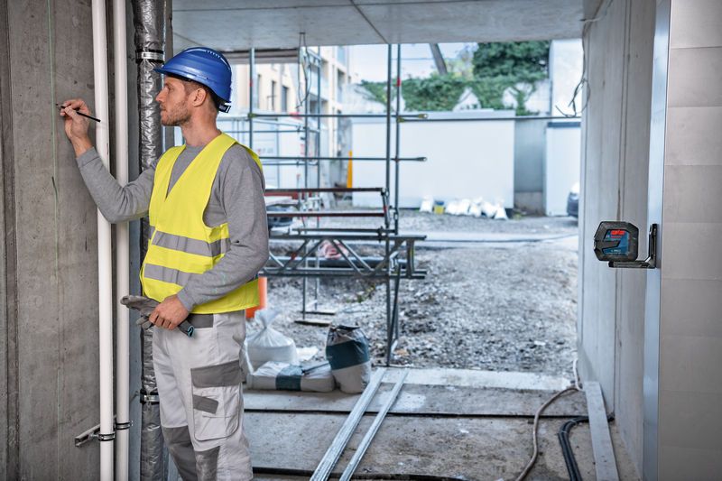 Uma pessoa que use equipamento de segurança marca uma parede, guiada por uma ferramenta de nivelamento a laser.