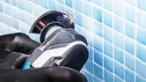 Uma pessoa que utilize equipamento de segurança utiliza uma ferramenta rotativa para cortar azulejos azuis na parede.