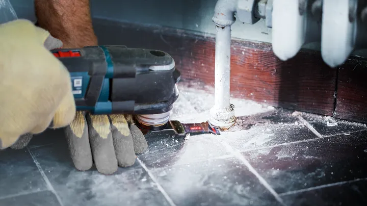 Uma pessoa que utiliza equipamento de segurança utiliza uma ferramenta elétrica para cortar o piso à volta de um cano.