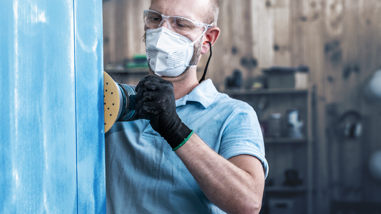 Uma pessoa que utilize equipamento de segurança lixa um painel azul com uma lixadora elétrica.