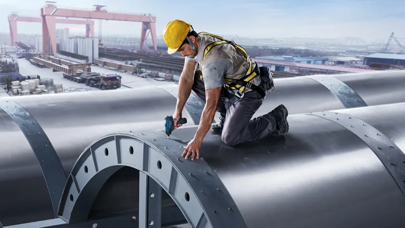 Uma pessoa que utiliza equipamento de segurança aperta parafusos numa grande estrutura metálica utilizando uma ferramenta elétrica.