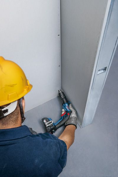 Uma pessoa que utiliza equipamento de segurança fixa uma parede de gesso com uma chave de fendas sem fios.