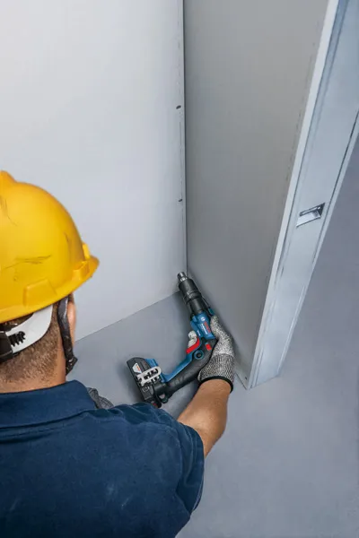 Uma pessoa que utiliza equipamento de segurança fixa uma parede de gesso com uma chave de fendas sem fios.