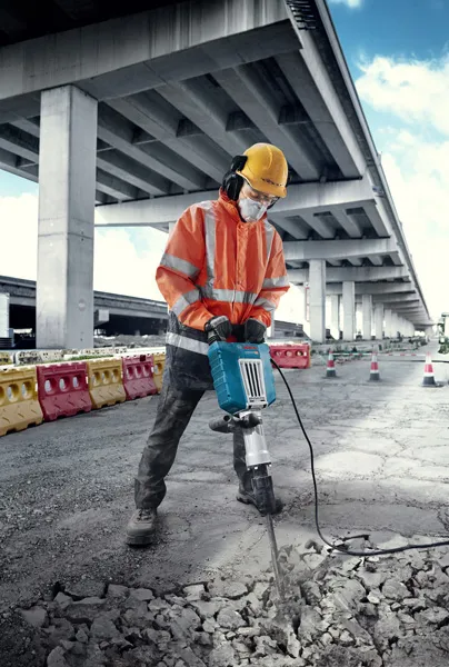 Uma pessoa que utiliza equipamento de segurança quebra betão com um grande disjuntor elétrico ao ar livre.