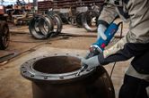 Uma pessoa que usa equipamento de segurança afia a borda de um grande cano de metal com uma retificadora reta.