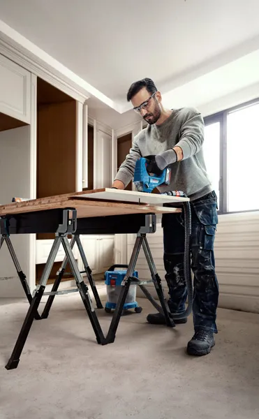 Uma pessoa que utiliza equipamento de segurança utiliza uma serra de tico-tico para cortar madeira numa bancada de trabalho.