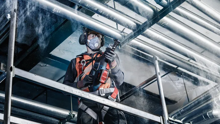 Pessoa que utiliza equipamento de segurança perfura um cano no teto de um estaleiro de construção.