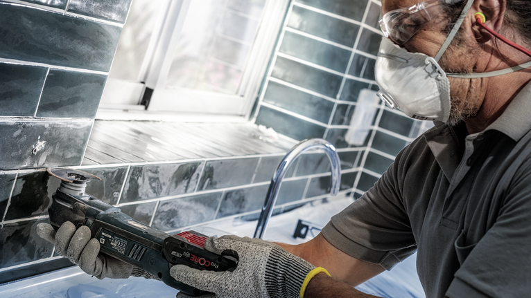 Uma pessoa que utiliza equipamento de segurança utiliza uma ferramenta elétrica para cortar azulejos perto de um lavatório.