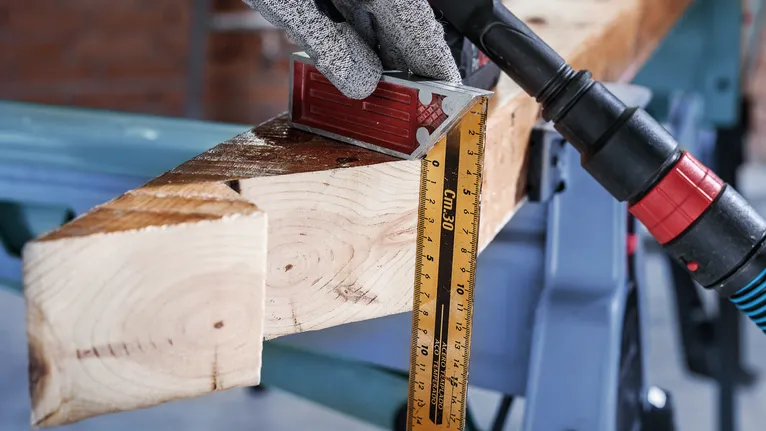 Uma pessoa que usa equipamento de segurança mede uma viga de madeira com uma régua e um esquadro.