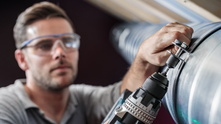 Uma pessoa que utiliza equipamento de segurança fixa um parafuso a uma conduta de metal com um berbequim elétrico.