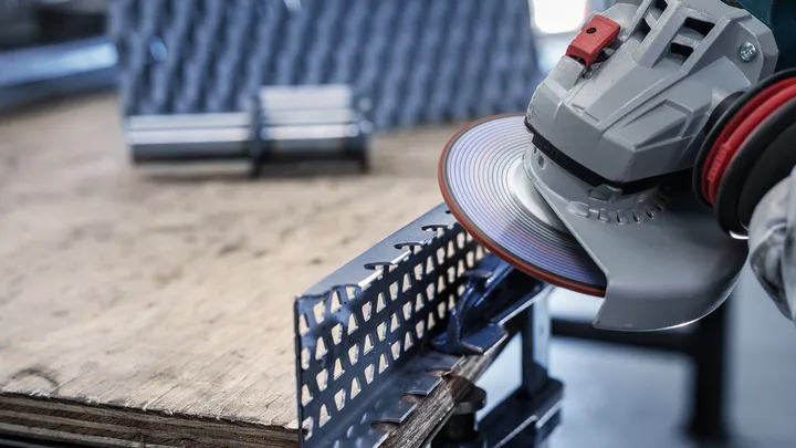 Uma pessoa que utilize equipamento de segurança afia uma barra angular de metal com uma ferramenta elétrica.