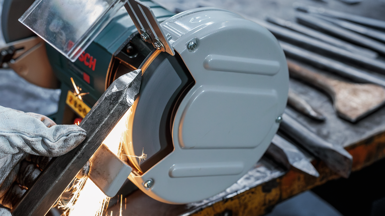 Uma pessoa que usa equipamento de segurança tritura metal numa rebarbadora de bancada, com faíscas a voar.