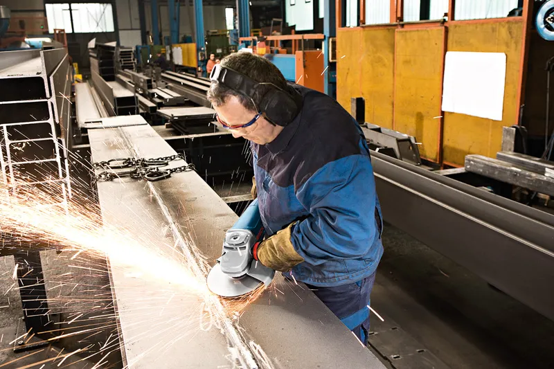 Uma pessoa que usa equipamento de segurança tritura uma viga metálica, produzindo faíscas.