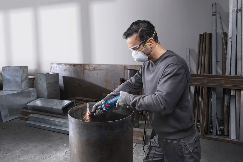 Uma pessoa que usa equipamento de segurança afia a borda de um barril de metal com uma máquina de retificação reta.