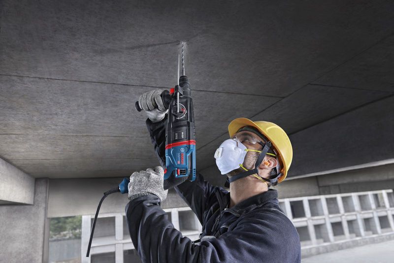 Uma pessoa que utilize equipamento de segurança perfura um teto de betão utilizando um martelo rotativo.