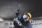 Uma pessoa que utilize equipamento de segurança perfura um teto de betão utilizando um martelo rotativo.