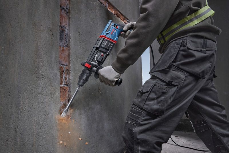 Uma pessoa que utiliza equipamento de segurança utiliza um martelo rotativo para cinzelar betão de uma parede.