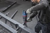 Uma pessoa que utilize equipamento de segurança perfura uma viga metálica com um martelo rotativo.